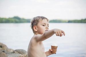 A cute blond boy appetizingly eats ice cream in the summer, sitting on the bank of the river. Cool off by the water. Funny facial expression. photo