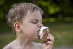 A cute blond boy appetizingly eats ice cream in the summer, sitting on the bank of the river. Cool off by the water. Funny facial expression. photo