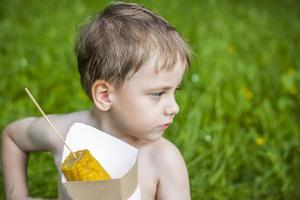 A cute blond boy appetizingly eats corn in the summer, sitting on the bank of the river on the juicy grass. Funny facial expression. photo