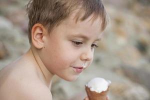 A cute blond boy appetizingly eats ice cream in the summer, sitting on the bank of the river. Cool off by the water. Funny facial expression. photo