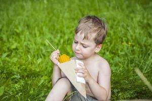 un lindo niño rubio come apetitosamente maíz en el verano, sentado en la orilla del río sobre la jugosa hierba. expresión facial divertida. foto