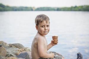 A cute blond boy appetizingly eats ice cream in the summer, sitting on the bank of the river. Cool off by the water. Funny facial expression. photo