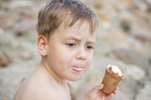 A cute blond boy appetizingly eats ice cream in the summer, sitting on the bank of the river. Cool off by the water. Funny facial expression. photo