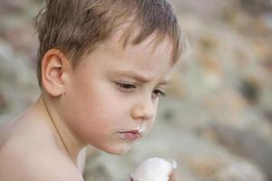 A cute blond boy appetizingly eats ice cream in the summer, sitting on the bank of the river. Cool off by the water. Funny facial expression. photo