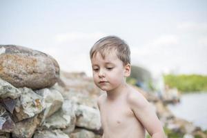 un lindo niño rubio experimenta la alegría de caminar en un día de verano junto al río, sentado en la jugosa hierba. expresión facial divertida. calor de verano, un niño sin camiseta. foto