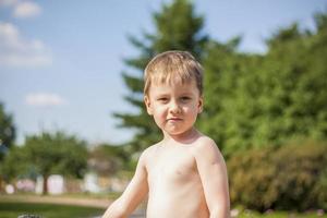 un lindo niño rubio experimenta la alegría de caminar en un día de verano junto al río, sentado en la jugosa hierba. expresión facial divertida. calor de verano, un niño sin camiseta. foto