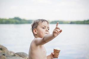 A cute blond boy appetizingly eats ice cream in the summer, sitting on the bank of the river. Cool off by the water. Funny facial expression. photo