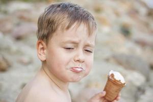 A cute blond boy appetizingly eats ice cream in the summer, sitting on the bank of the river. Cool off by the water. Funny facial expression. photo