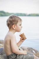 A cute blond boy appetizingly eats ice cream in the summer, sitting on the bank of the river. Cool off by the water. Funny facial expression. photo