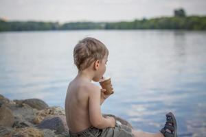 A cute blond boy appetizingly eats ice cream in the summer, sitting on the bank of the river. Cool off by the water. Funny facial expression. photo