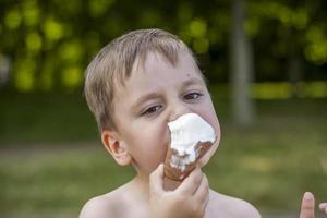 A cute blond boy appetizingly eats ice cream in the summer, sitting on the bank of the river. Cool off by the water. Funny facial expression. photo