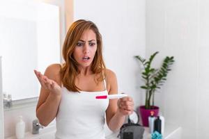 mujer sorprendida mirando la línea de control en la prueba de embarazo. chica joven sorprendida con un embarazo no deseado mirando la prueba en el baño. triste joven sosteniendo una prueba de embarazo sintiéndose desesperanzada foto