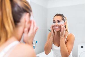 mujer joven después de la ducha mirándose en el espejo mimándose a sí misma aplicándose mascarilla facial use un producto de belleza anti-envejecimiento sintiéndose feliz, cuidado de la piel, concepto de rutina matutina diaria foto