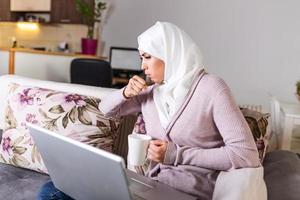 joven musulmana que tiene un problema de cogida en casa. joven musulmana enferma sentada en un sofá, sofá y usando una laptop en la sala de estar en casa, enfermedad y frío foto