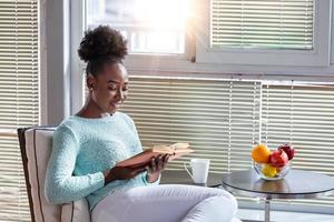 sofá acogedor y una chica hermosa, leyendo un libro, conceptos de hogar y comodidad, lugar para el texto. hermosa joven negra leyendo un libro cerca de la ventana en casa foto