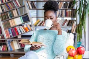 Young woman with a book and cup of hot drink. Pretty afro-american girl with reading a book sitting in a home library with bookshelves in the back and enjoying her coffee. photo