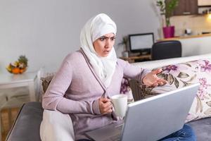 Portrait of muslim woman looks angry while working on laptop. Young Arabic woman having video call via laptop at home. photo