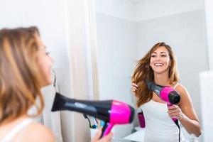 Happy woman drying her hair in bathroom. Photo taken in mirror. Beautiful young woman is using a hair dryer and smiling while looking into the mirror in bathroom