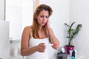 Shocked woman looking at control line on pregnancy test. Shocked young girl with unwanted pregnancy looking the test in the bathroom. sad young woman holding pregnancy test feeling hopeless photo