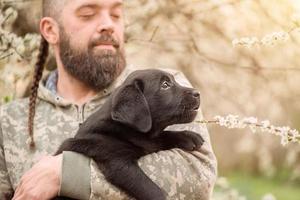 A small black Labrador retriever puppy. A dog in the arms of a bearded man. photo