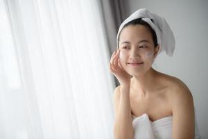 Happy young Asian woman applying face lotions while wearing a towel and touching her face. Daily makeup and skincare photo