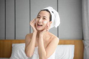 Happy young Asian woman applying face lotions while wearing a towel and touching her face. Daily makeup and skincare photo