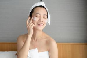 Happy young Asian woman applying face lotions while wearing a towel and touching her face. Daily makeup and skincare photo