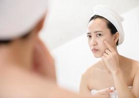 Problem skin. Concerned young asian women popping pimple on cheek while standing near mirror in bathroom. young asian women with acne photo