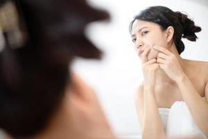 Problem skin. Concerned young asian women popping pimple on cheek while standing near mirror in bathroom. young asian women with acne photo