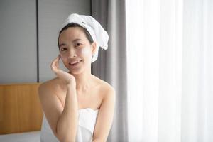 Happy young Asian woman applying face lotions while wearing a towel and touching her face. Daily makeup and skincare photo