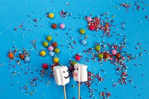 marshmallows on wooden skewers with color sweets on blue background. holiday concept. sweets in the form of people photo