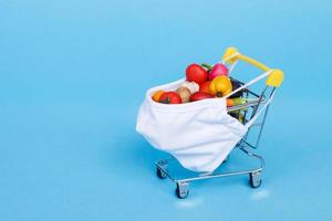 carrito de compras con máscara médica con frutas y verduras sobre un fondo azul. compras en línea en concepto de cuarentena. foto