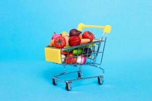 Metal shopping cart with fruits and vegetables on a blue background. Toy miniature shopping trolley photo