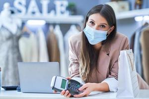 Young woman cashier in clothes store with terminal in mask photo