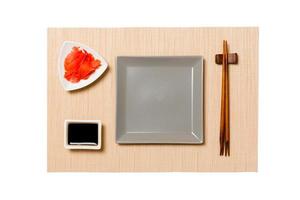 Empty gray square plate with chopsticks for sushi, ginger and soy sauce on brown sushi mat background. Top view with copy space for you design photo