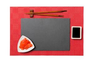 placa de pizarra negra rectangular vacía con palillos para sushi, jengibre y salsa de soja sobre fondo de sushi de alfombra roja. vista superior con espacio de copia para su diseño foto