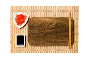 Empty rectangular brown wooden plate with chopsticks for sushi, ginger and soy sauce on yellow bamboo mat background. Top view with copy space for you design photo