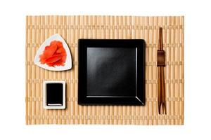 Empty black square plate with chopsticks for sushi, ginger and soy sauce on yellow bamboo mat background. Top view with copy space for you design photo