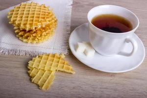 desayuno con pila de gofres en servilleta y trozos de gofre con taza blanca de té negro sobre superficie de madera foto