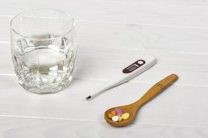 Pills with thermometer and glass of water on white wooden table. Medical concept photo