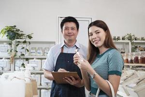 Asian male shopkeeper and female customer smile and look at camera at natural products in reusable containers at refill store, zero-waste groceries, organic retail, and sustainable shopping lifestyle. photo