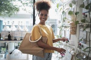 A Black female customer fills liquid soap into recycling jar from reusable containers in a zero-waste and refill store, smiles, and looks at camera, happy shopping in an environment-friendly shop. photo