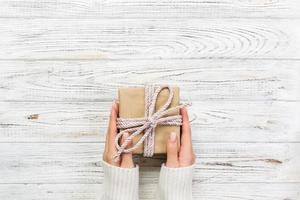 Woman hands give wrapped valentine or other holiday handmade present in paper with pink ribbon. Present box, decoration of gift on white wooden table, top view with copy space photo