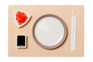 Empty round white plate with chopsticks for sushi, ginger and soy sauce on brown sushi mat background. Top view with copy space for you design photo