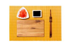 Empty rectangular brown wooden plate with chopsticks for sushi, ginger and soy sauce on yellow bamboo mat background. Top view with copy space for you design photo