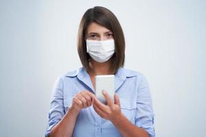 Attractive woman wearing protective mask holding smartphone isolated over white background photo
