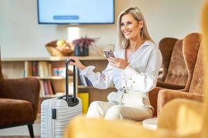 Businesswoman with luggage in modern hotel lobby using smartphone photo