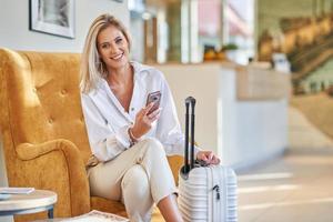 Businesswoman with luggage in modern hotel lobby using smartphone photo