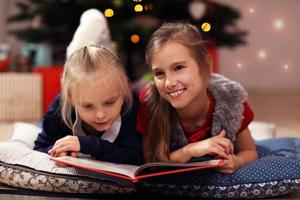 dos lindas hermanitas leyendo un libro de cuentos juntas bajo el árbol de navidad foto