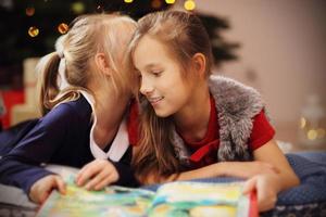 dos lindas hermanitas leyendo un libro de cuentos juntas bajo el árbol de navidad foto